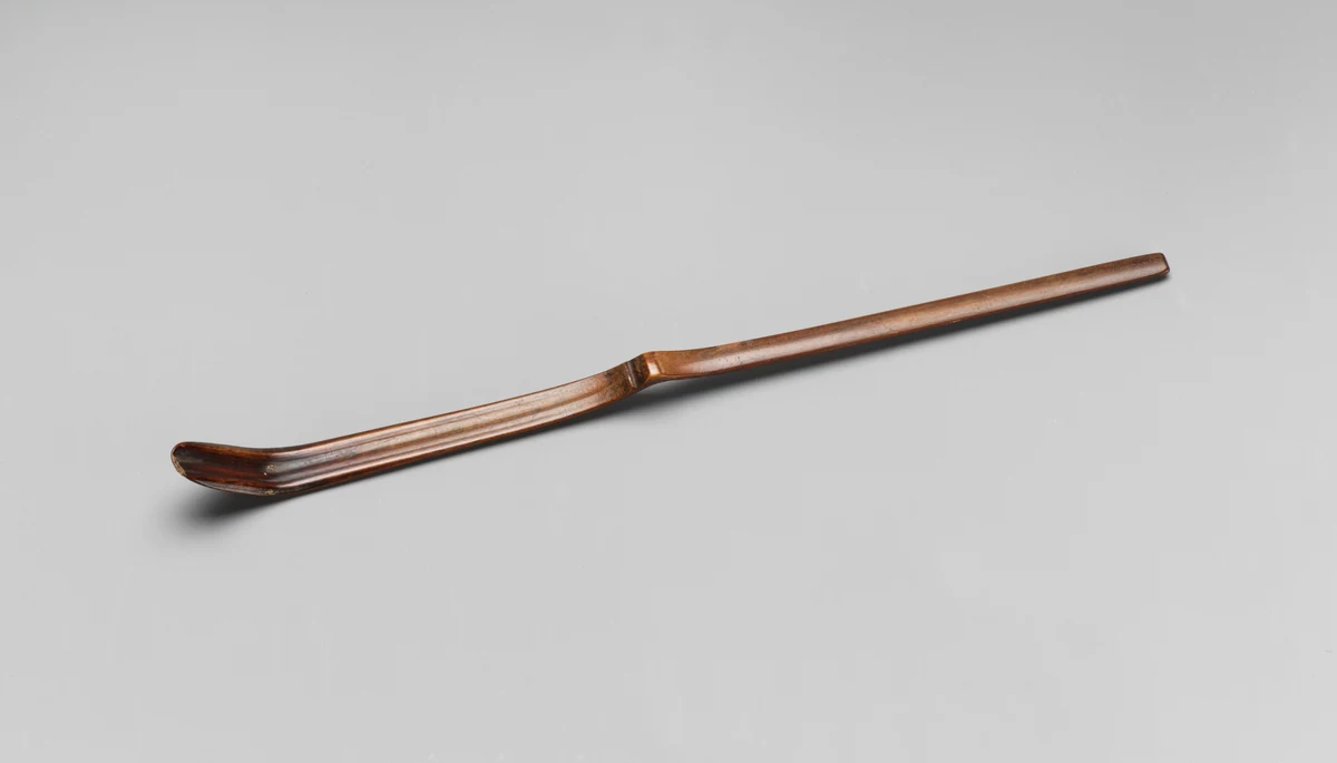A slender bamboo tea scoop with a bent bowl rests against a gray background.