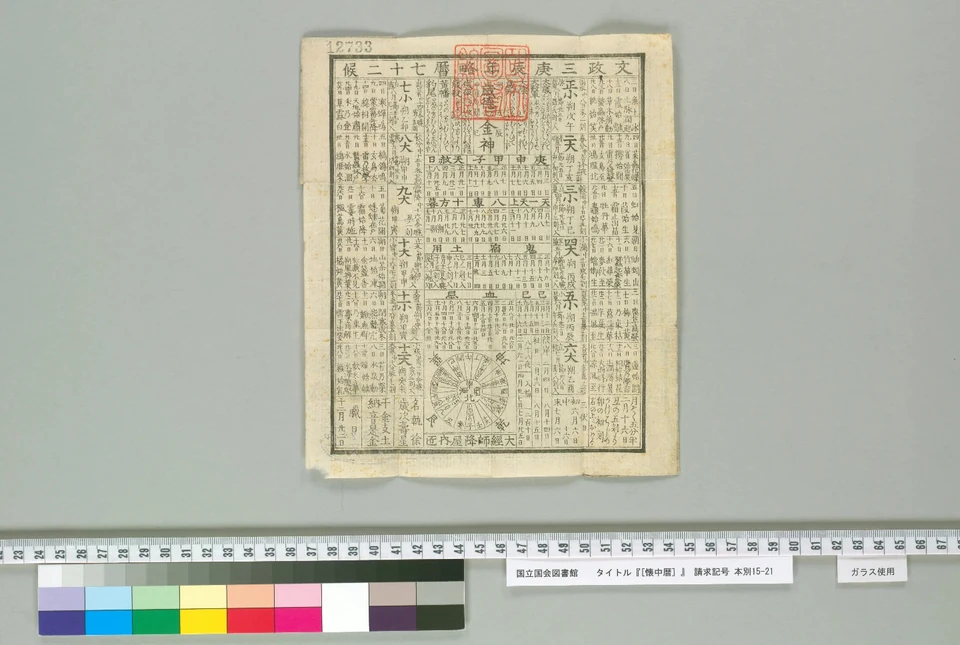 An unfolded pocket calendar contains dense vertical writing, visible fold lines and a red stamp; modern recording ruler and colour scale sit outside the sheet.