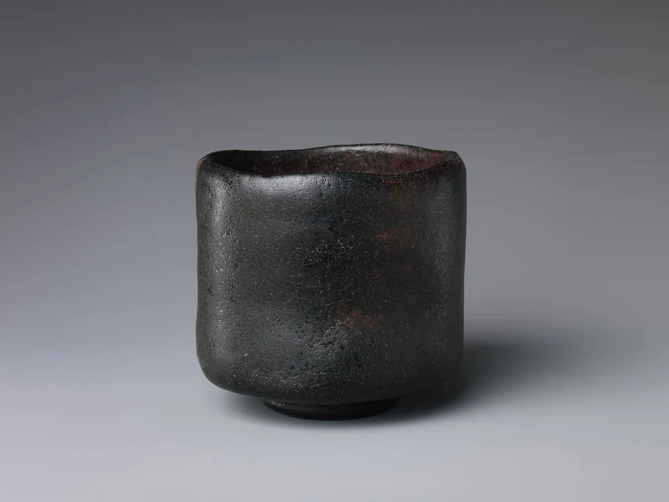 An early-seventeenth-century black Raku bowl has an uneven lip, nearly cylindrical body, varied dark glaze and a small foot.