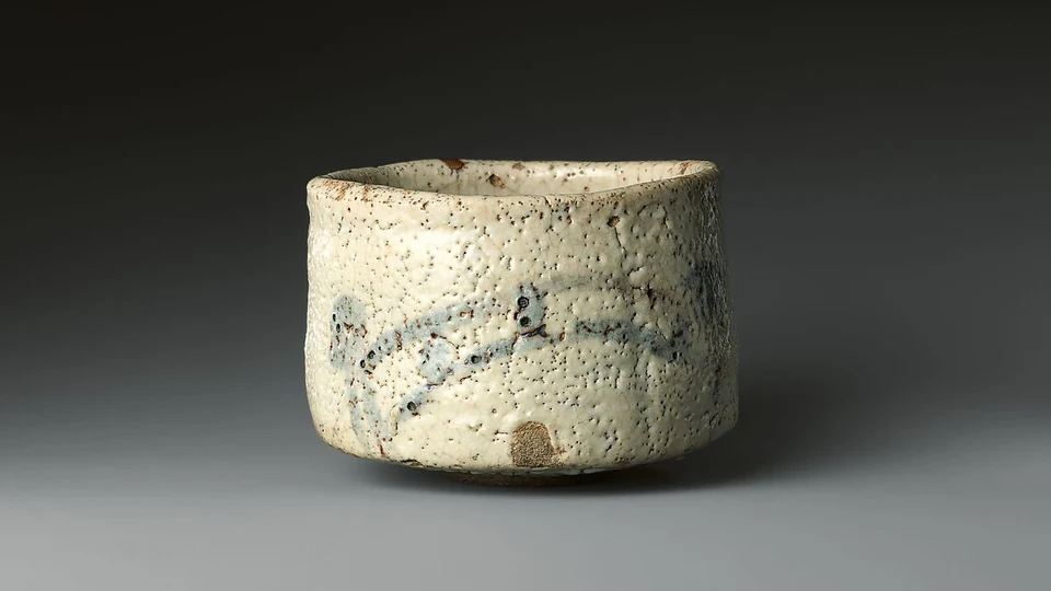 A pale Shino tea bowl has an uneven rim, pitted white glaze, faint brown bridge strokes and an unglazed spot near its lower front.