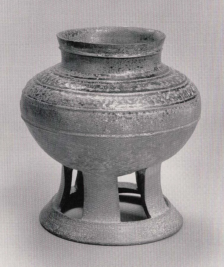 A grey Sue-ware cup stands on a tall flaring foot pierced by rectangular openings. Its raised mouth has no surviving lid in this photograph.